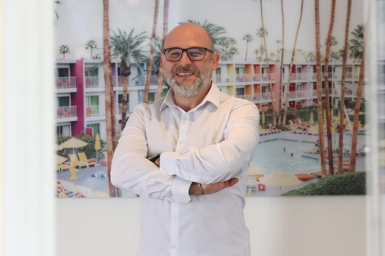 Hakan Duganci, Vetriebsleiter der paxconnect GmbH, steht in einem weißen Hemd mit verschränkten Armen vor einem Bild, das ein farbenfrohes Hotel mit Palmen und Pool zeigt.