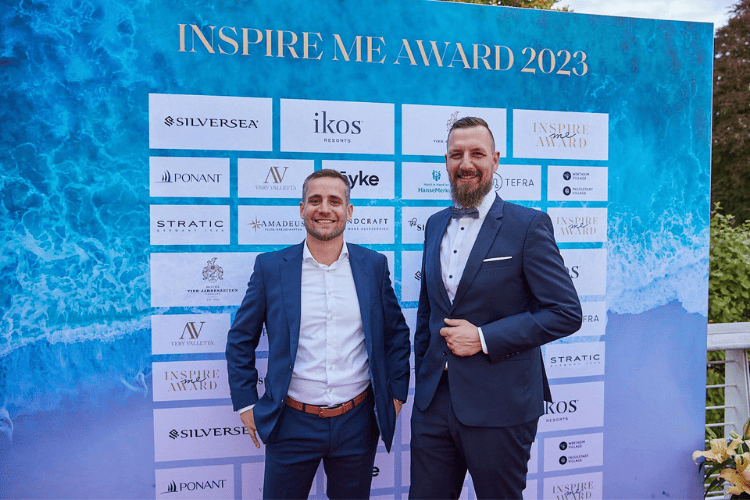 Zwei Männer in eleganter Abendgarderobe posieren vor einer Sponsorenwand beim 'Inspire Me Award 2023'. Der Hintergrund zeigt das Logo des Events sowie die Logos verschiedener Sponsoren, darunter Silversea, ikos Resorts und Ponant.